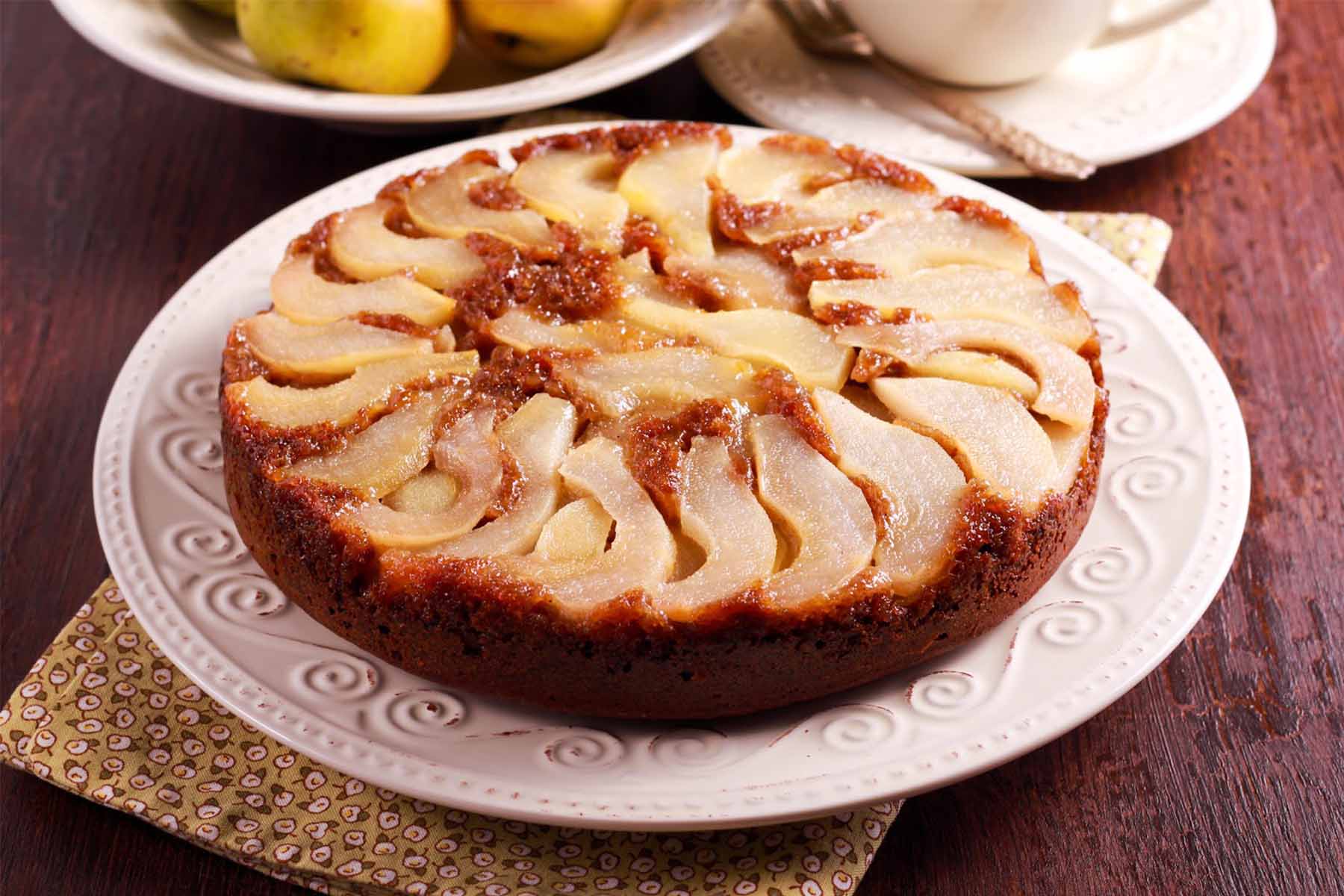 A beautifully presented pear upside-down cake on a decorative white plate, featuring a caramelized pear topping, surrounded by bowls of fruit and a cup in the background.