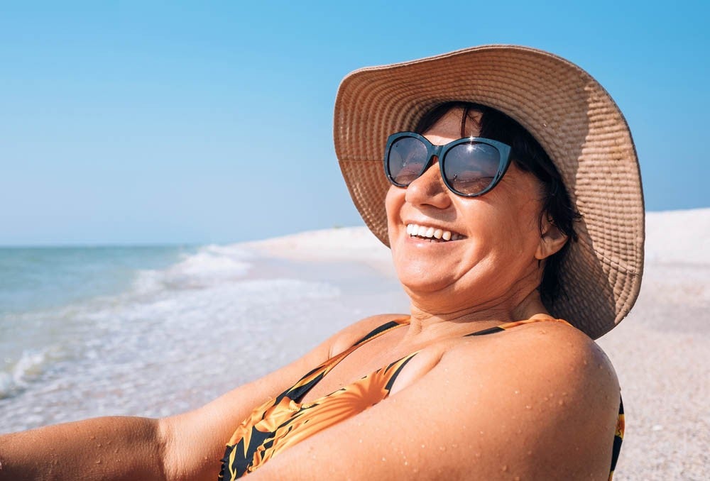Happy smiling elderly woman in sunglasses and big hat sun bathing on seaside