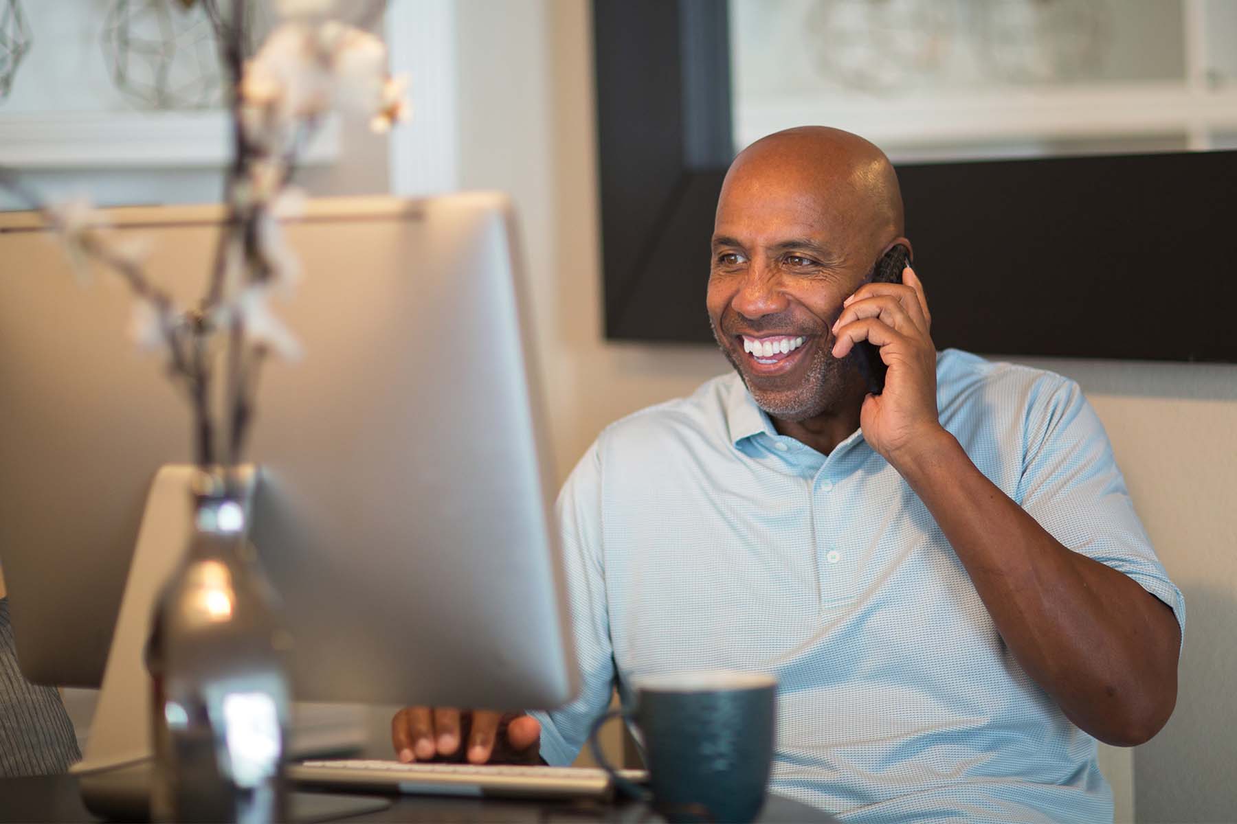 Man smiling while working at a computer and talking on the phone in a home office.