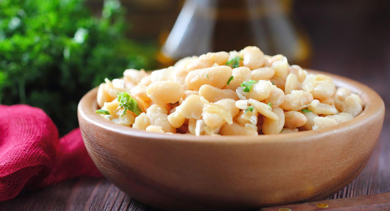 Bowl of cannellini beans garnished with herbs sits on a wooden table. A red napkin and fresh greens create a fresh, inviting atmosphere.