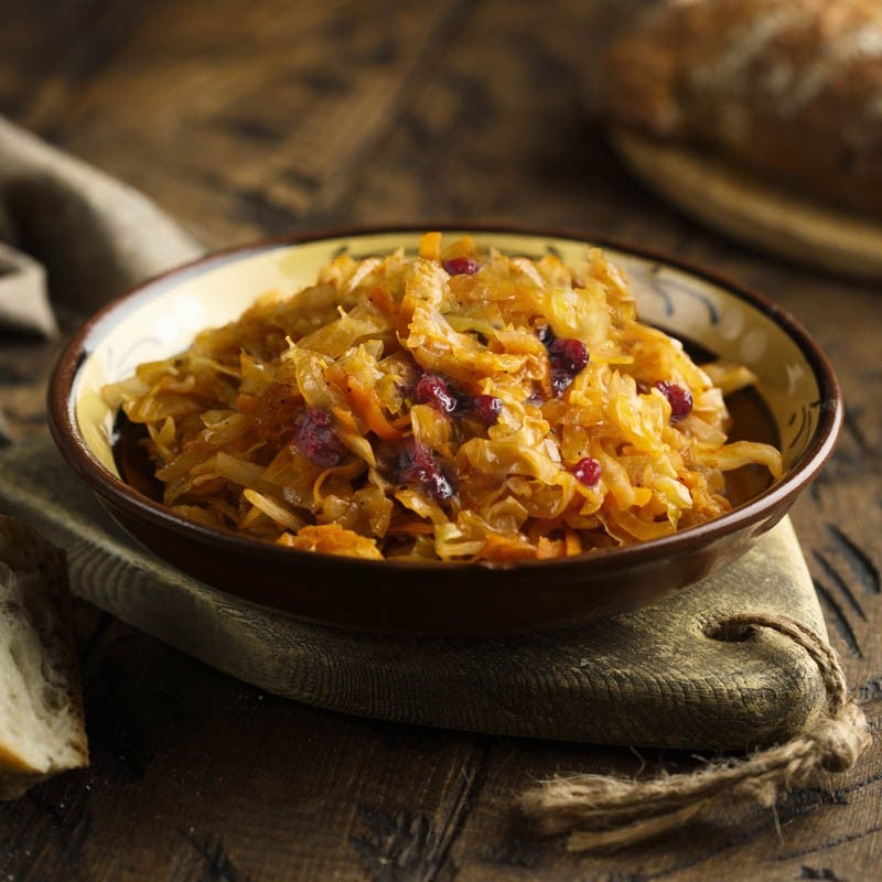 Un cuenco rústico lleno de chucrut de color dorado con trozos rojos, sobre una superficie de madera texturizada con una rebanada de pan al fondo