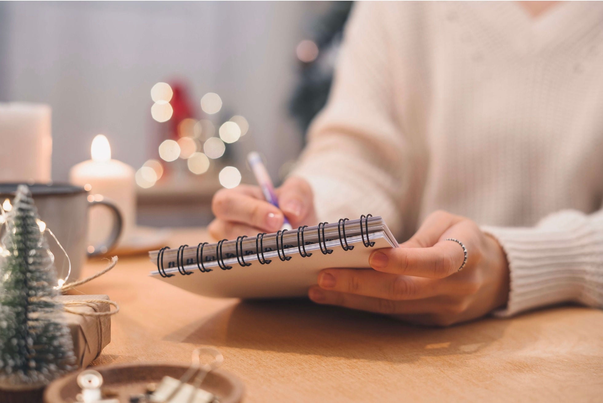 Woman sitting at a table with holiday decorations in the background, writing with a pen and notepad.
