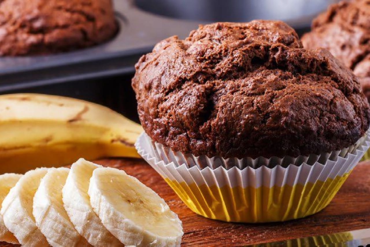 A chocolate and banana muffin resting on a cutting board next to banana slices with an unpeeled banana in the background.