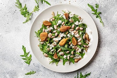 Plato de ensalada con hojas verdes, queso feta desmenuzado, frijoles blancos y higos secos sobre una superficie clara