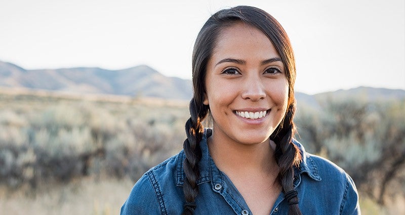 Smiling woman with braided hair standing outdoors in a natural landscape.