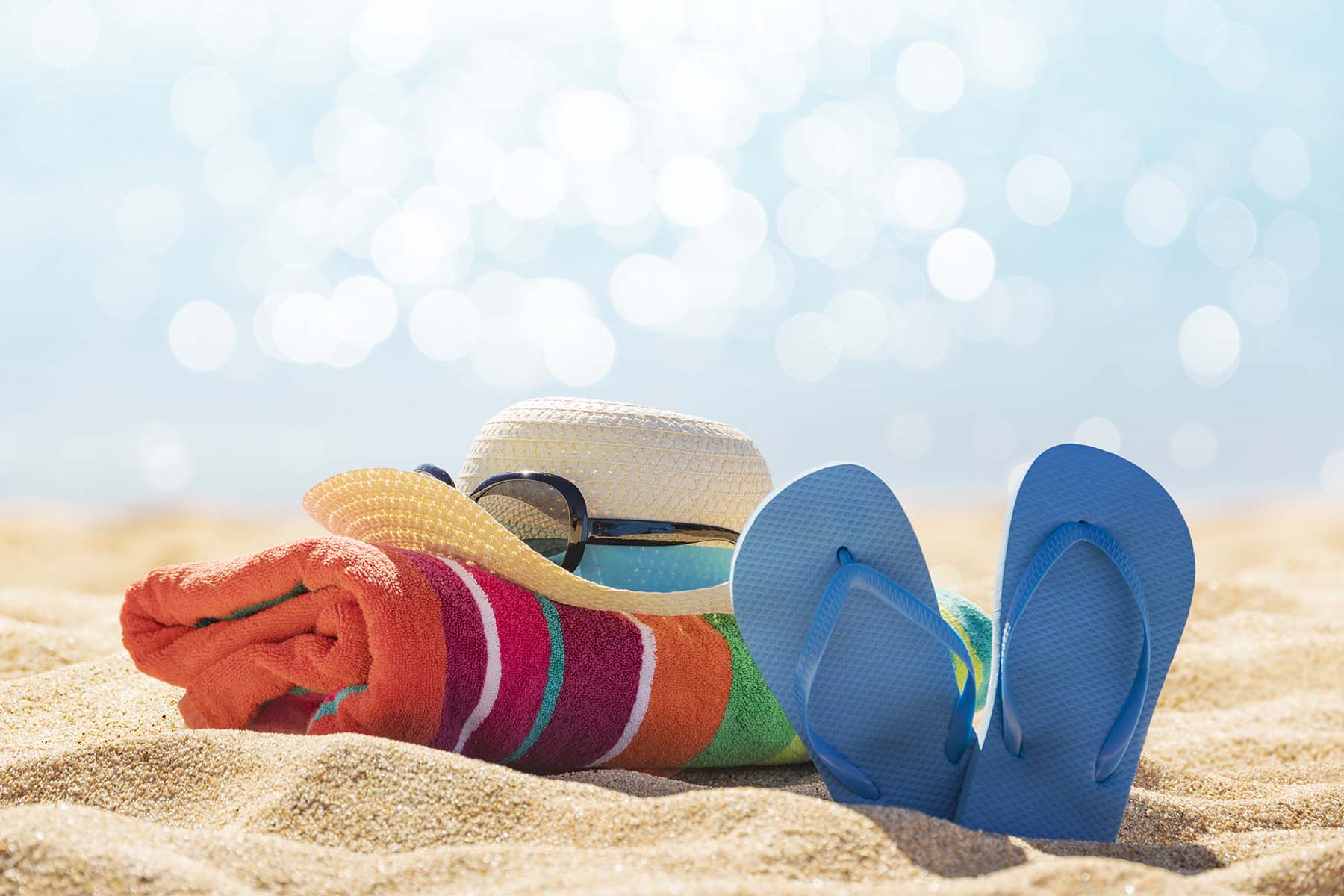 Beach essentials including a straw hat, sunglasses, striped towel, and blue flip-flops on the sand.