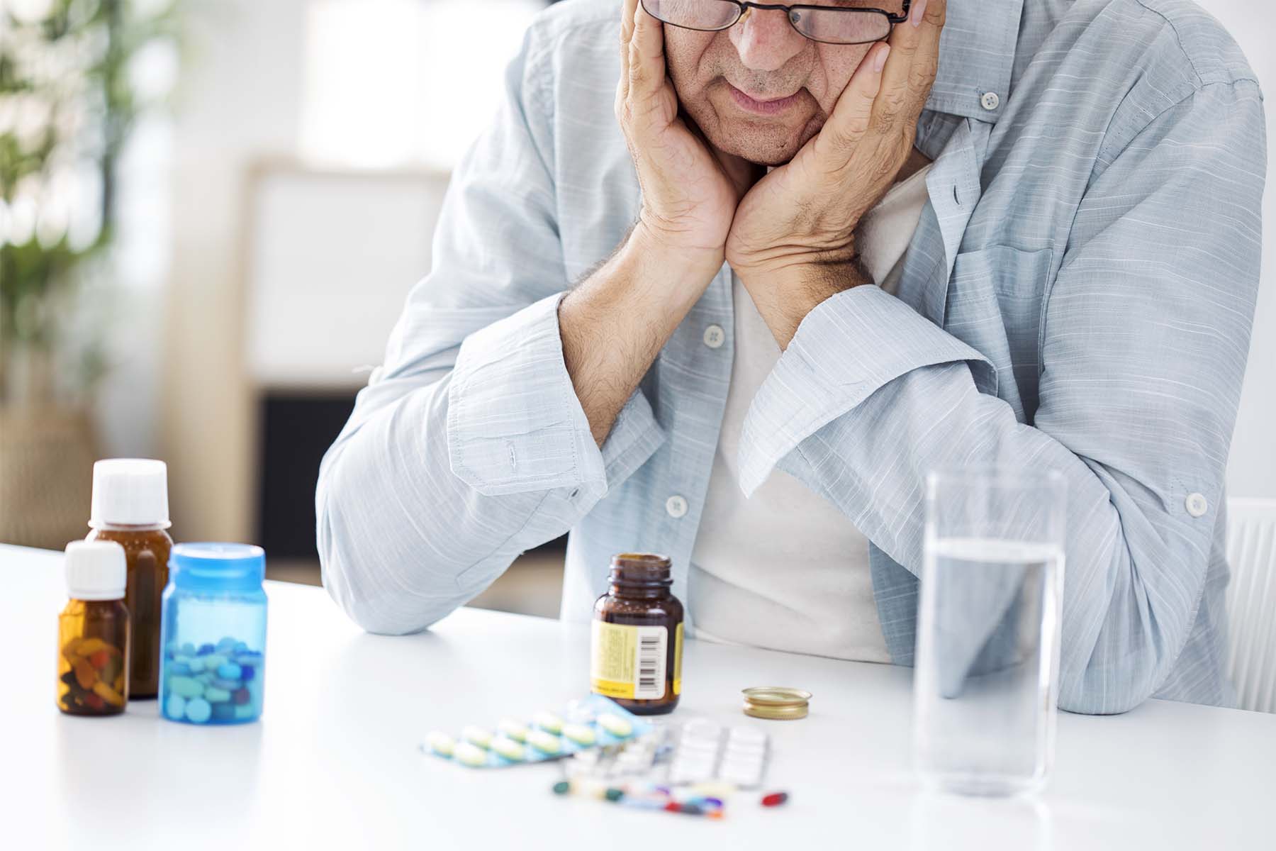Older man with glasses holding his face in his hands, looking at various medication bottles and pills on a table.