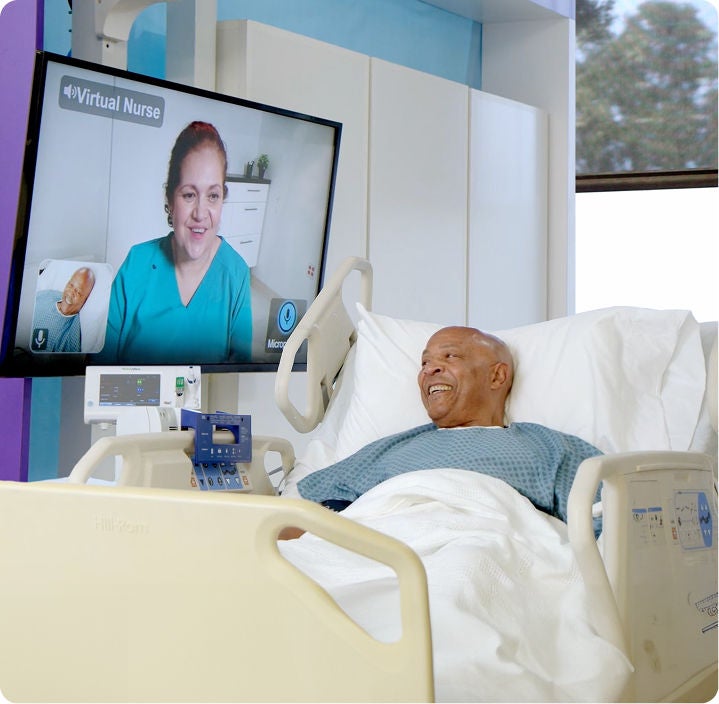 A patient in a hospital bed smiling while speaking with a virtual healthcare professional on a screen.