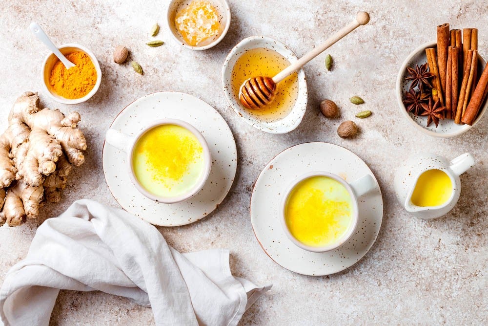 Flat lay of turmeric golden milk with ingredients including ginger, honey, cinnamon sticks, star anise, and cardamom on a light stone background