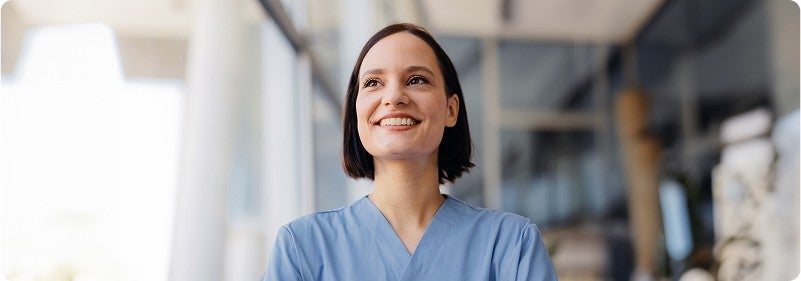 Confident female healthcare professional in blue scrubs smiling outdoors in front of a modern medical facility.
