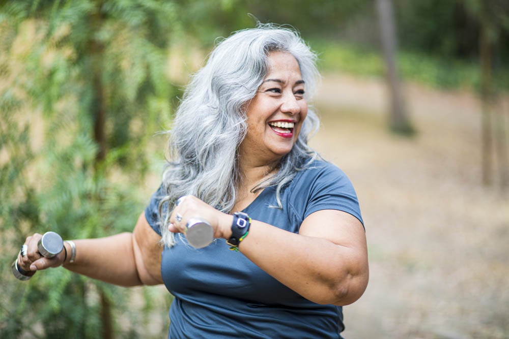 Smiling woman with gray hair exercises outdoors using small dumbbells, wearing a blue shirt and watch, surrounded by trees.