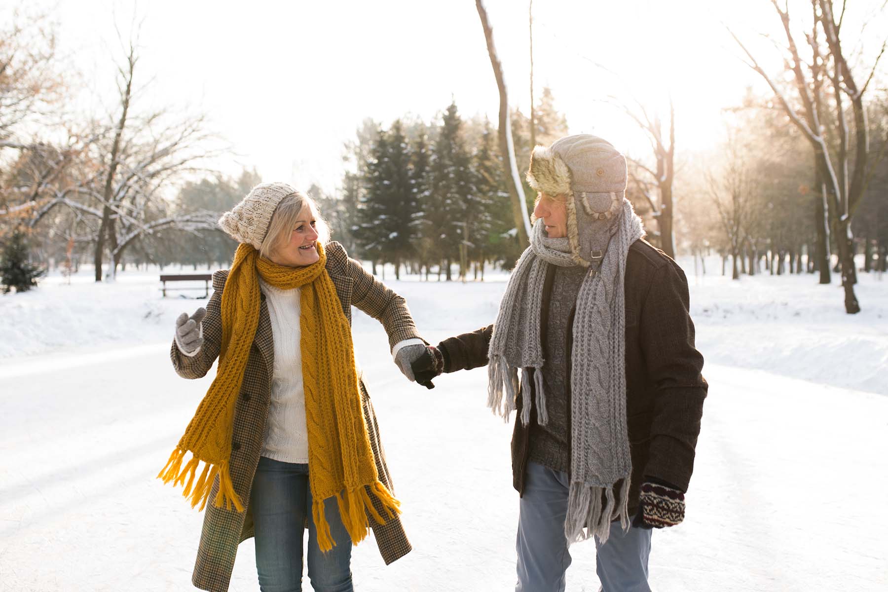 Older couple walking hand in hand on a snowy path, bundled up in winter coats, scarves, and hats, with the sun shining through the trees.