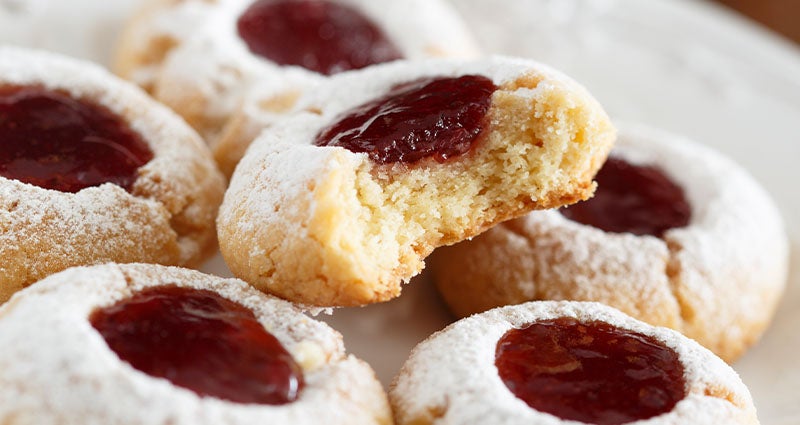 Jam-filled cookies dusted with powdered sugar, one with a bite taken out, arranged on a plate.