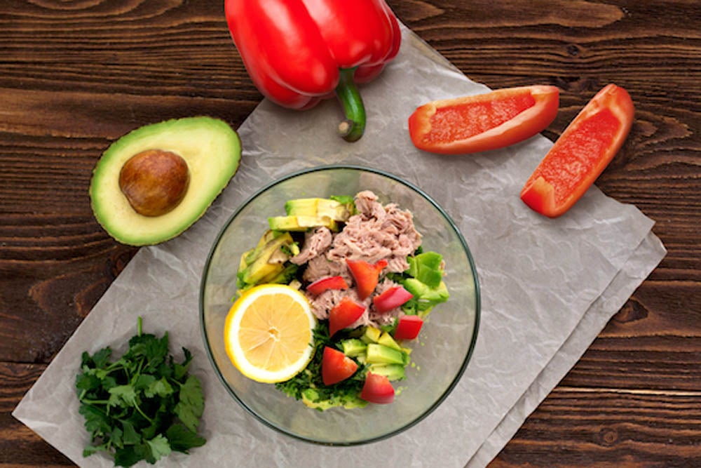 A bowl with tuna, avocado, tomatoes, and lemon slices on a table. Surrounding items: cilantro, a halved avocado, and red bell pepper pieces.