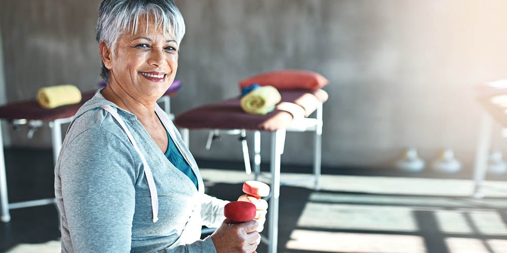 Una mujer mayor sonriente con el cabello gris corto, vestida con una sudadera con capucha gris, sosteniendo mancuernas rojas en un entorno de fitness, con colchonetas y toallas visibles en el fondo.