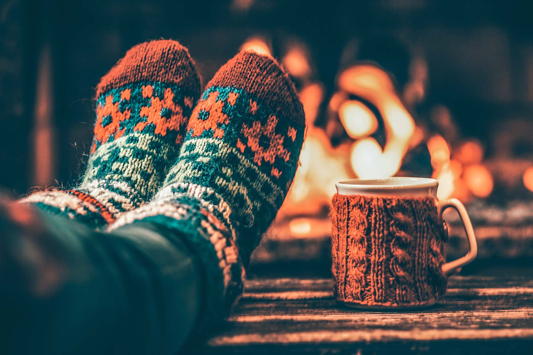 Feet in cozy patterned socks resting by a fireplace with a mug of hot drink nearby.