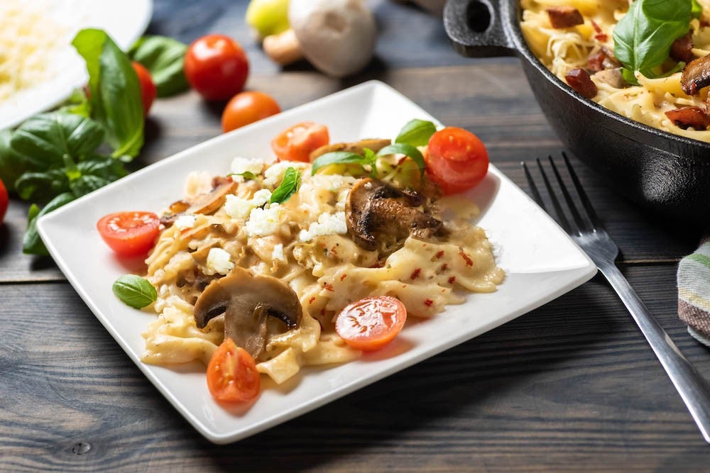 Plato cuadrado de pasta con pollo en cubos, tomates cherry y hojas de albahaca fresca, con una olla negra al fondo sobre una mesa de madera