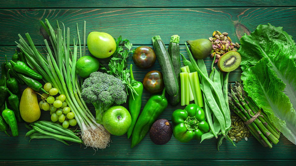 Mesa verde con frutas y verduras verdes alineadas, como brócoli, palta, kiwi, espárragos, lima y pistachos, vista desde arriba.