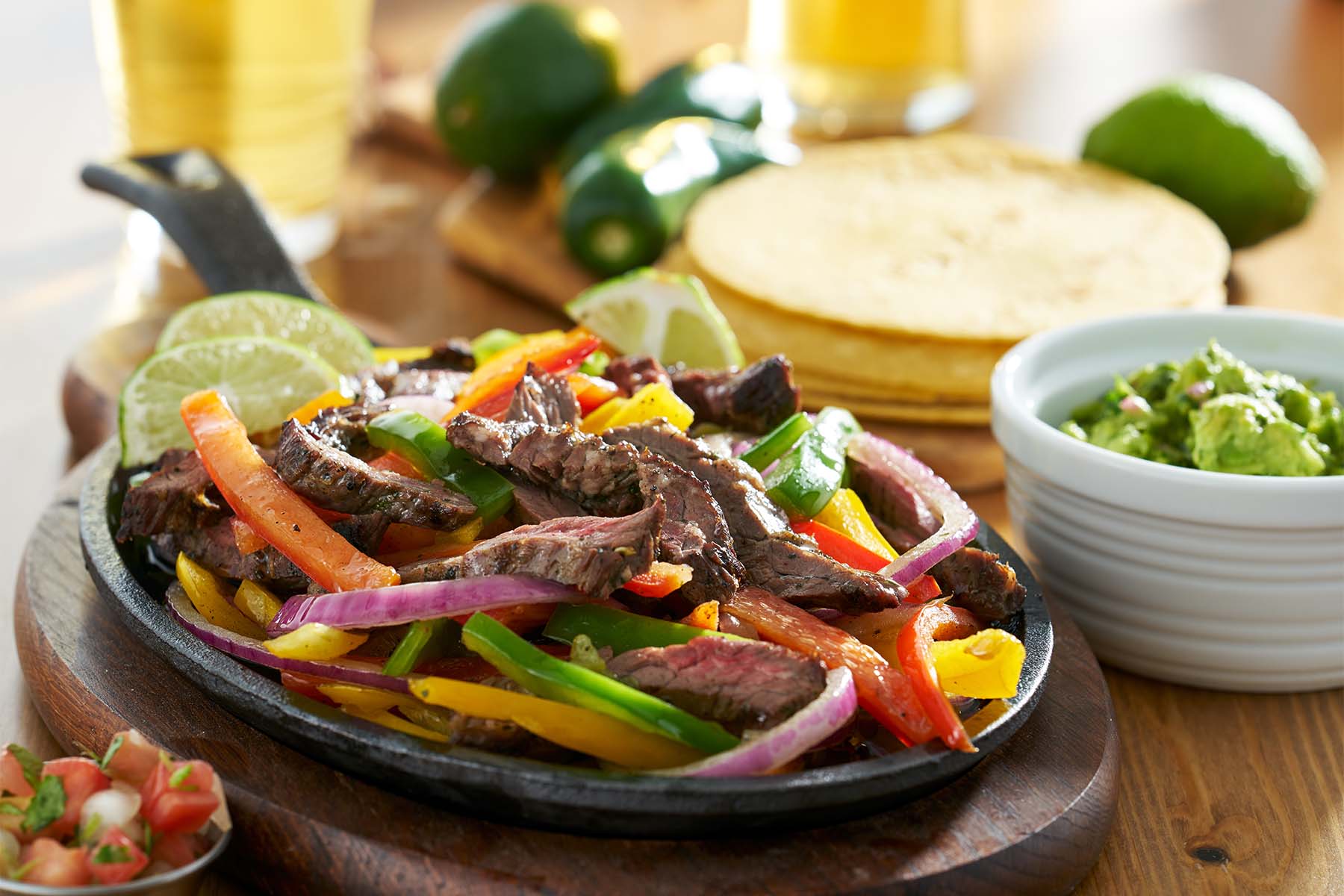 A sizzling platter of fajitas with sliced beef, colorful peppers, and onions, garnished with lime wedges, accompanied by a bowl of guacamole and tortillas on a wooden table.