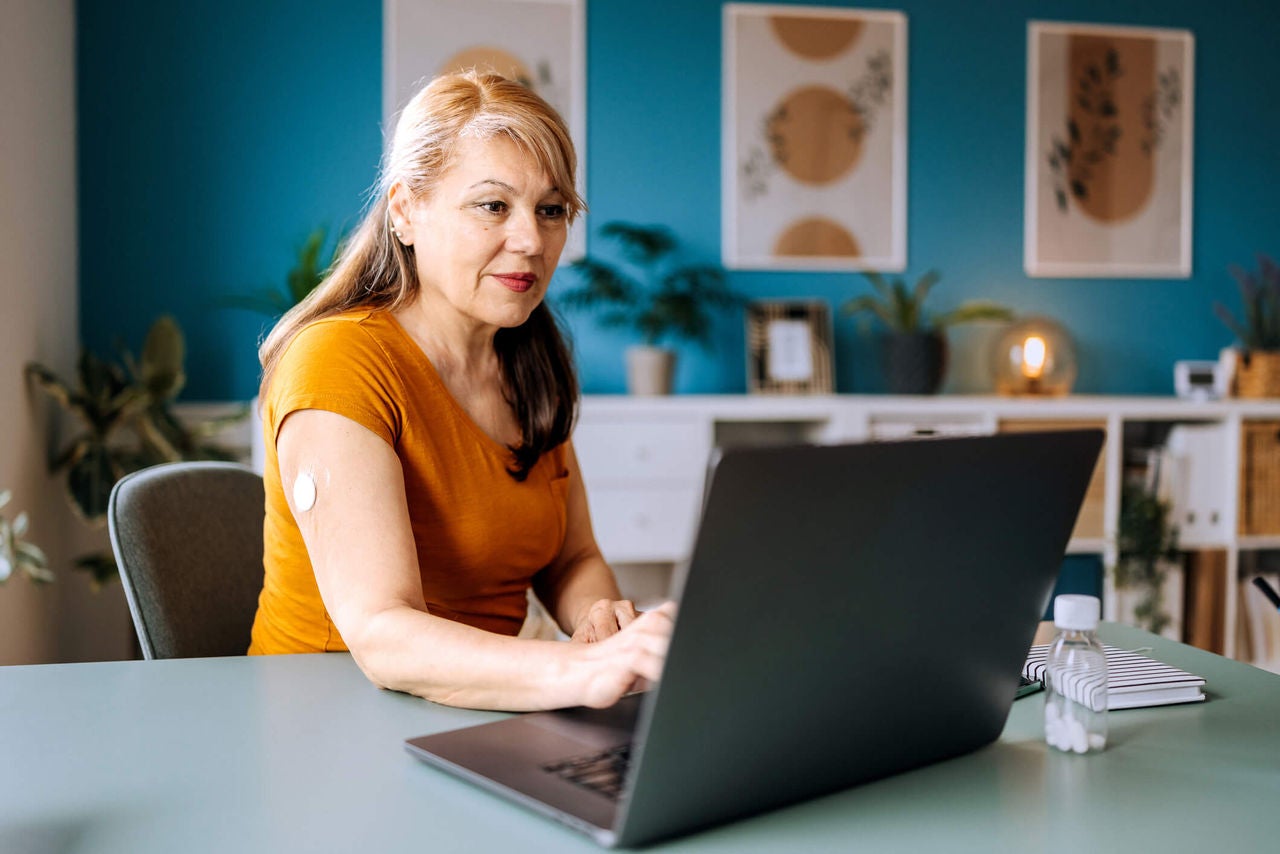 Woman wearing a cgm monitor seated at a table using a laptop computer in a brightly lit room.