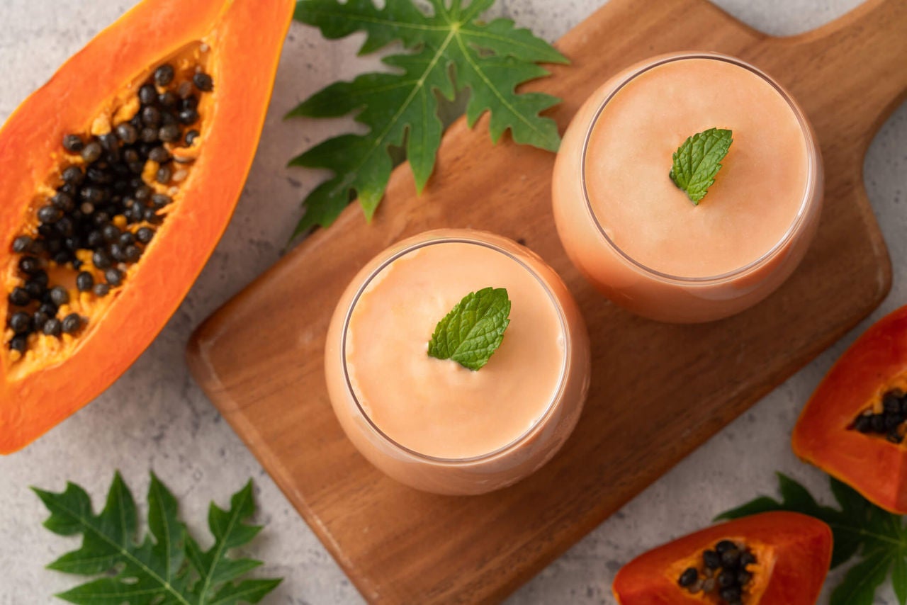 Two orange smoothies topped with mint leaves on a wooden board, with papaya segments on the side.