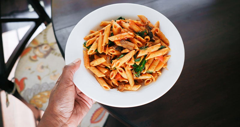 A hand holding a white bowl filled with penne pasta tossed in a red sauce, garnished with green herbs, set against a blurred background of a dining area.