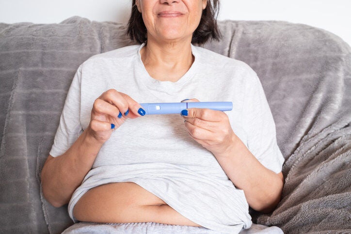 Woman sitting on a couch holding an injector pen while smiling, her stomach partially exposed. 
