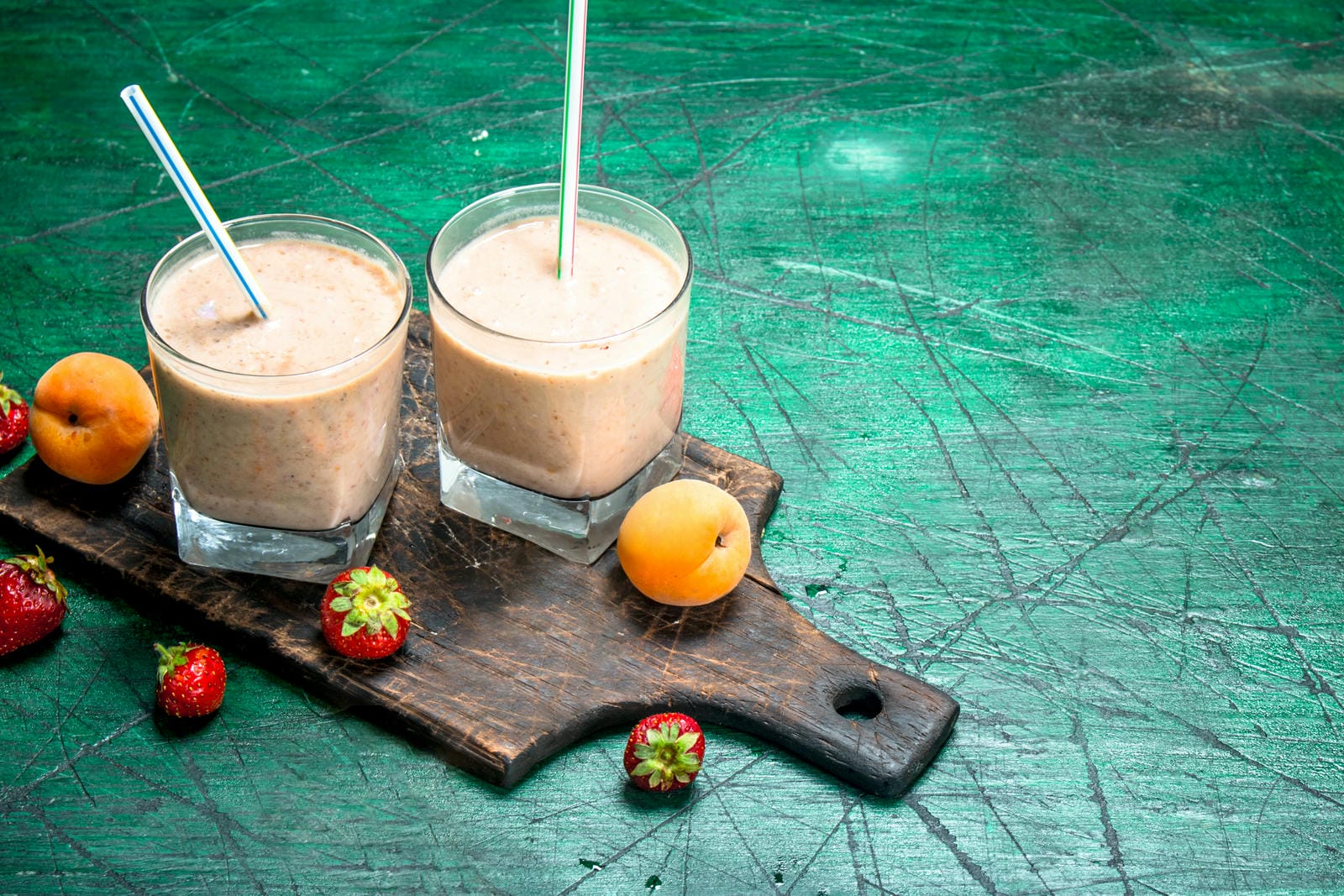 Dos vasos de batido de frutas con pajillas, colocados sobre una tabla de madera, acompañados de albaricoques y fresas sobre una superficie verde envejecida.