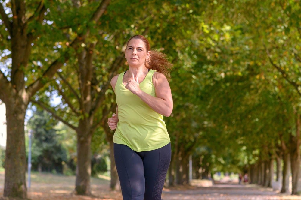 Mujer de mediana edad en forma corriendo por una avenida arbolada a través de un parque, acercándose a la cámara en un concepto de salud y fitness o estilo de vida activo