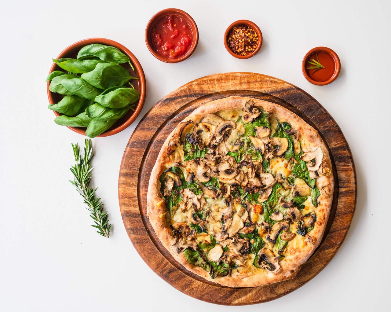 Pizza topped with veggies on a wooden tray, surrounded by healthy ingredients in small bowls. 