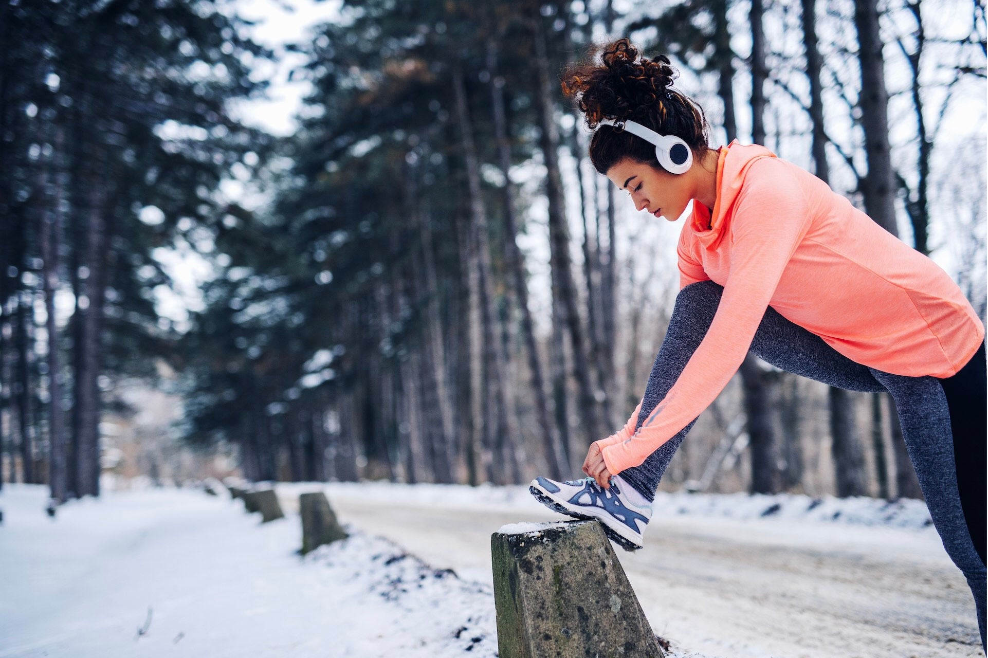 Woman wearing athletic clothing tying her running shoe on a snowy path. 