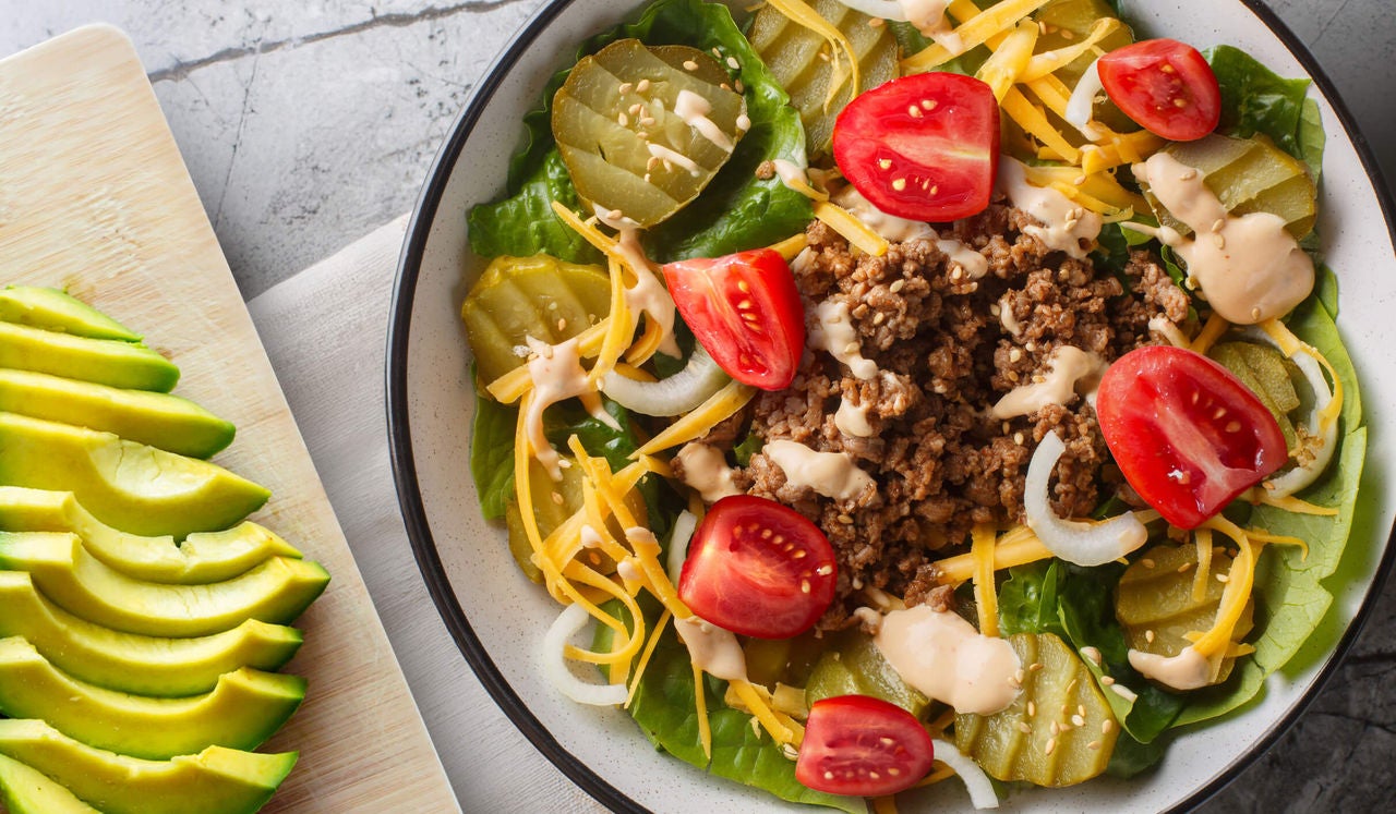 Burger salad with ground meat, tomatoes, pickles, onions and dressing with sliced avocado on the side.