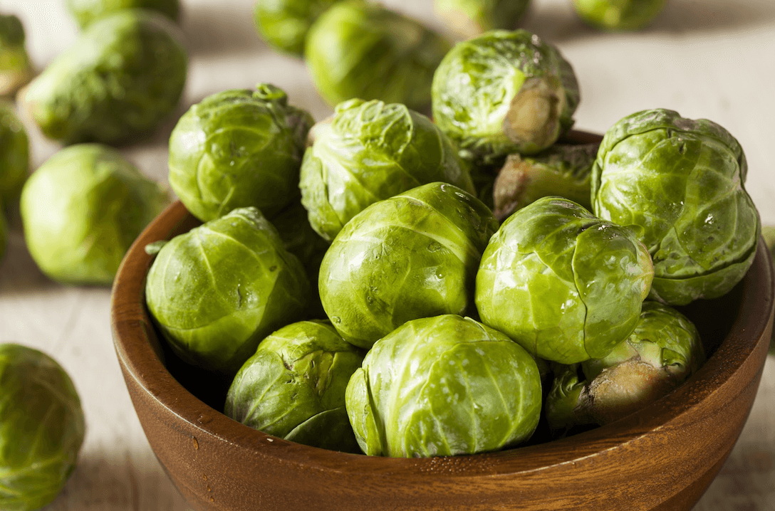 A wooden bowl filled with fresh green Brussels sprouts on a light surface, with a few loose sprouts scattered around.