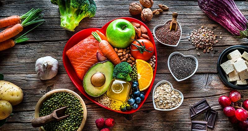 An assortment of fresh foods arranged in a heart-shaped container, including salmon, fruits, vegetables, nuts, and seeds, displayed on a rustic wooden surface.