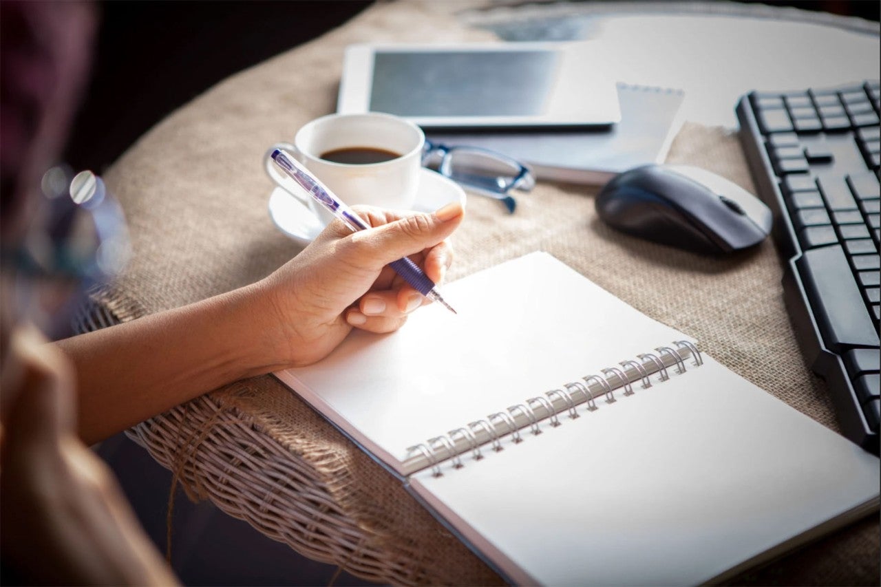 Close-up of a person writing in a spiral notebook with a pen, accompanied by a cup of coffee, glasses, a tablet, and a computer keyboard and mouse on a textured burlap surface, suggesting a workspace setup.