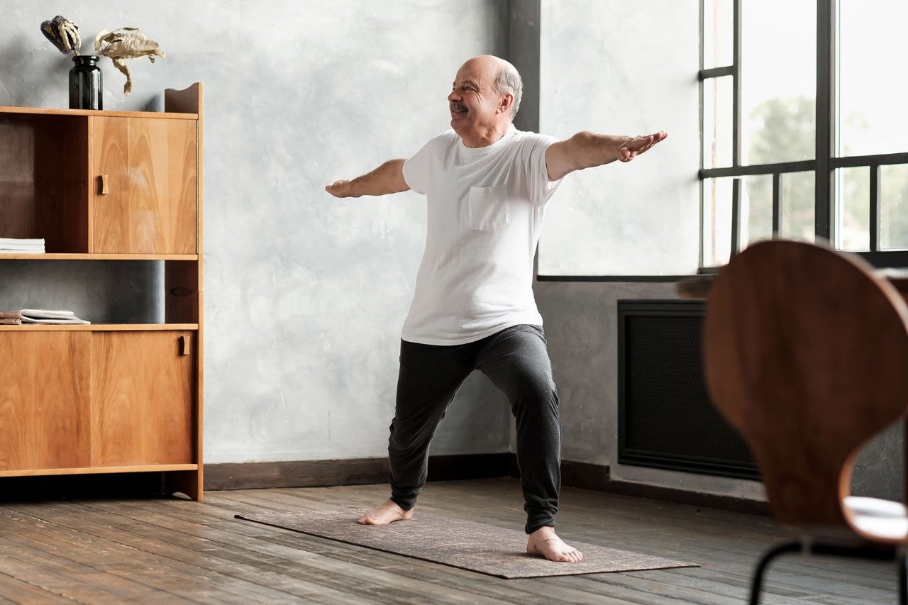 Older man practicing yoga in a bright room, standing in warrior pose with arms extended.