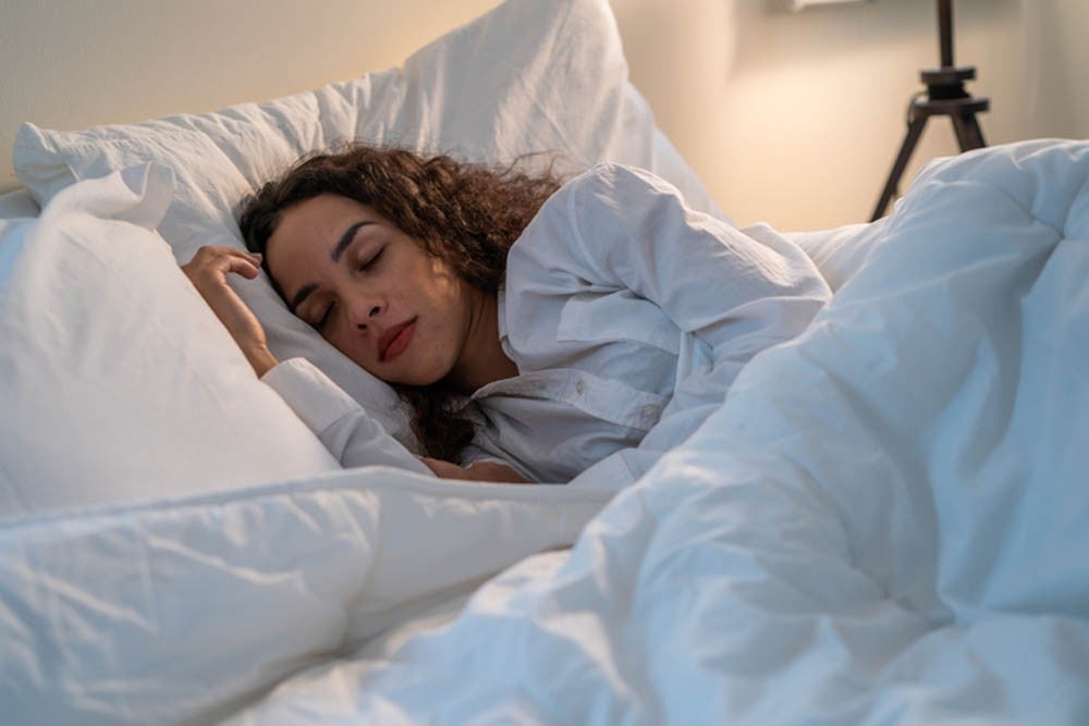 A person with curly hair sleeping peacefully in a bed, wearing a white shirt, surrounded by white bedding and pillows, under soft lighting.