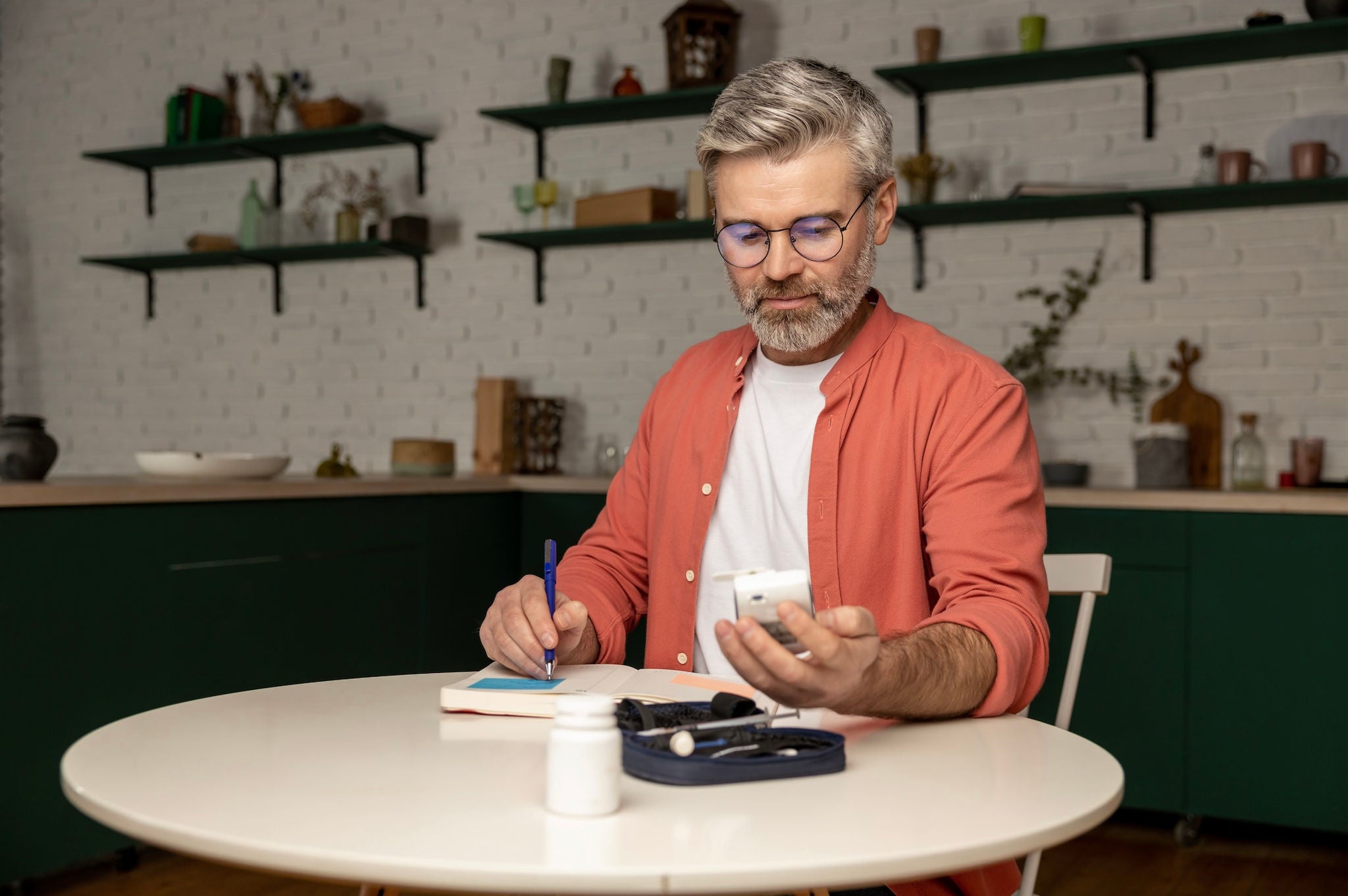 Man sitting at table with a blood glucose test kit, reading from a blood sugar meter while writing in a notebook.