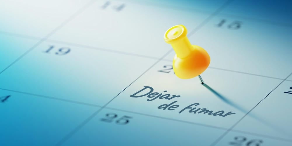Una chincheta amarilla sobre un cuadrado de un calendario azul que tiene escrito en su interior la frase "dejar de fumar".