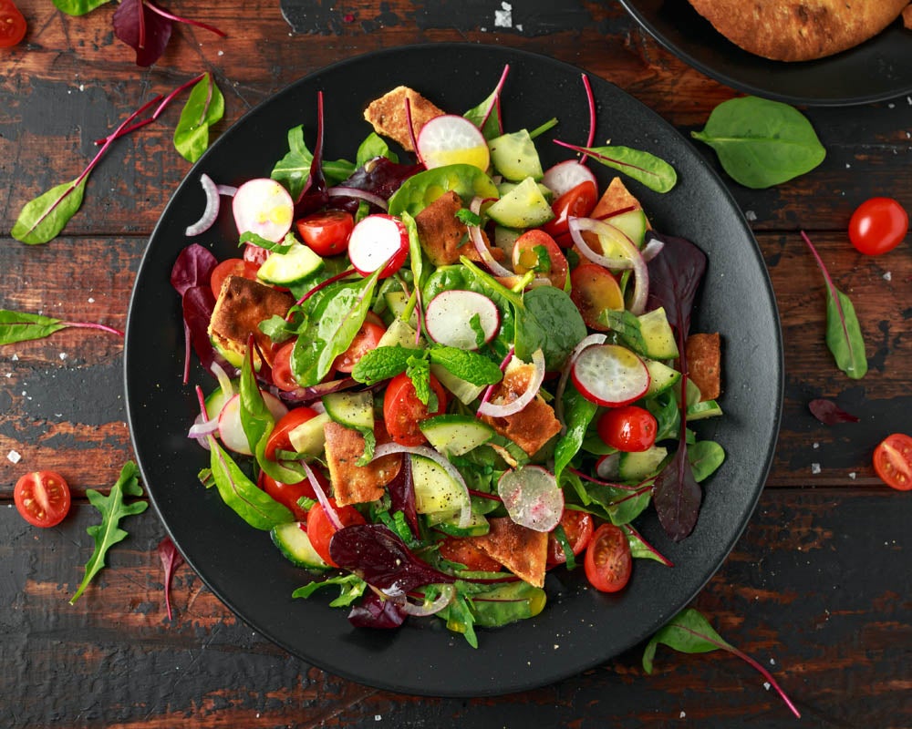 Ensalada vibrante con mezcla de hojas verdes, tomates cherry, pepino, rábanos y trozos de pan pita, servida en un plato negro sobre un fondo rústico de madera