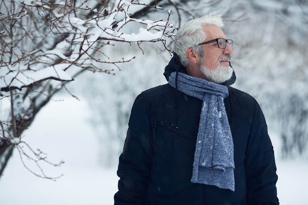 Active grandpa concept. Portrait of handome mature man in trendy winter clothing and fashionable glasses standing near bushes, over snow park background. Close up. Text-space.  Outdoor shot