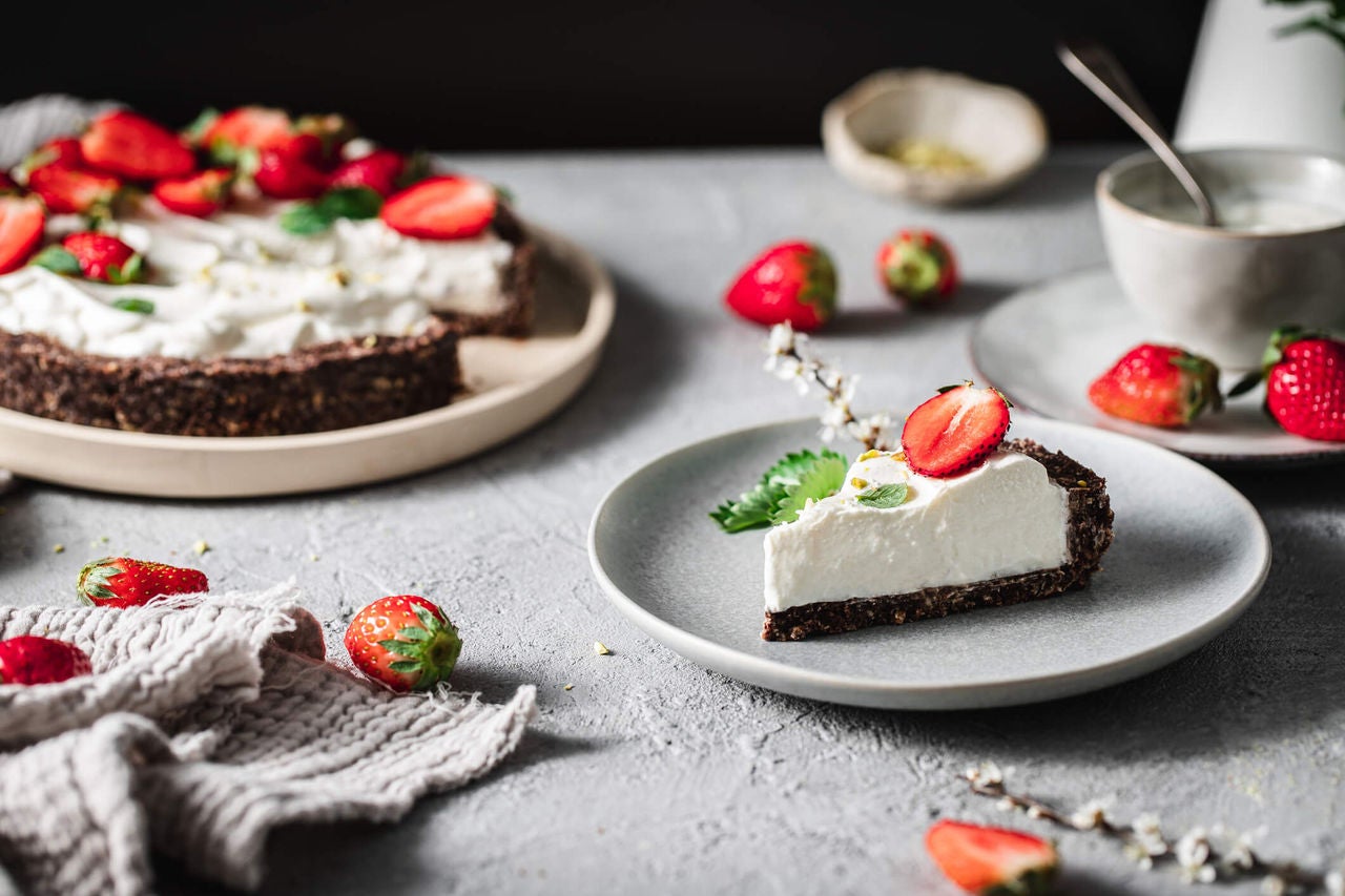 Slice of white yogurt tart with vegan crust on a plate with full tart in the background, topped with sliced and full strawberries.