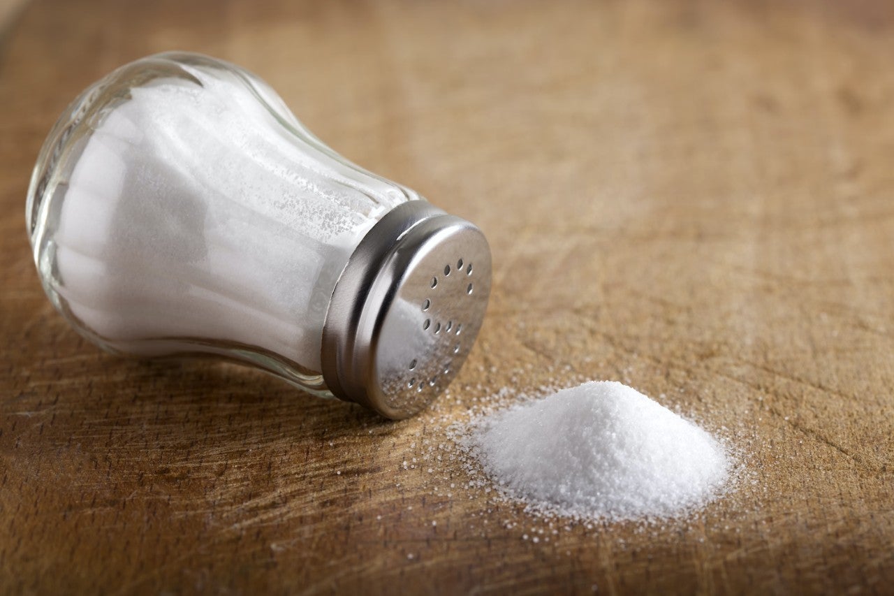 A tilted salt shaker lies on its side on a wooden surface, with a small mound of spilled salt next to it.