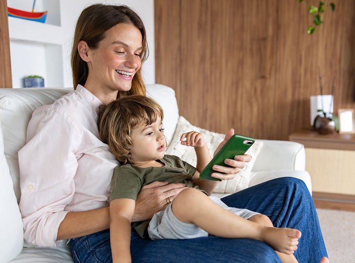 Mother sitting on a couch with her young child, smiling while looking at a smartphone together at home.