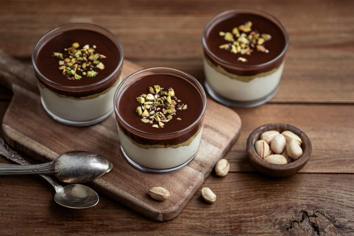 Three glass cups containing a chocolate pistachio yogurt parfait on a wooden table with spoons and a bowl of pistachios.