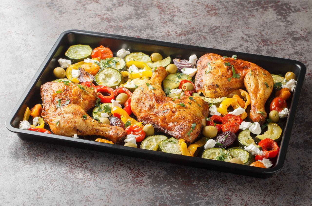 Sheet pan with cooked marinated chicken, assorted vegetables and crumbled feta cheese on a counter surface.