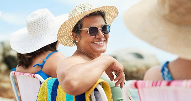 Smiling woman in a sun hat and sunglasses sitting on a beach chair with friends.