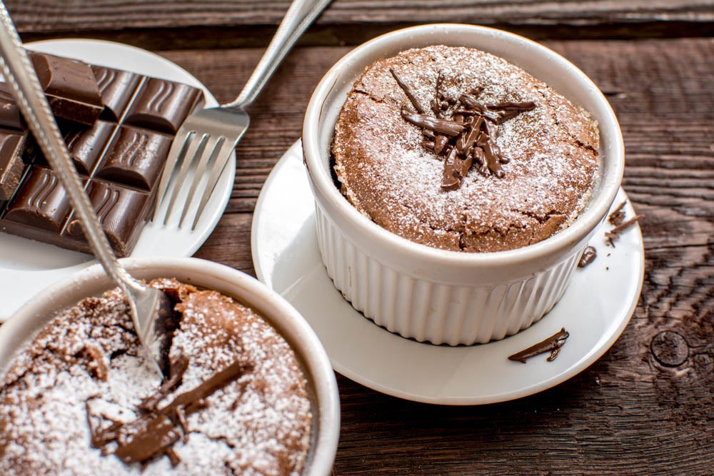 Dos soufflés de chocolate espolvoreados con azúcar glas, servidos en ramequines, junto a un plato con trozos de chocolate y una cuchara