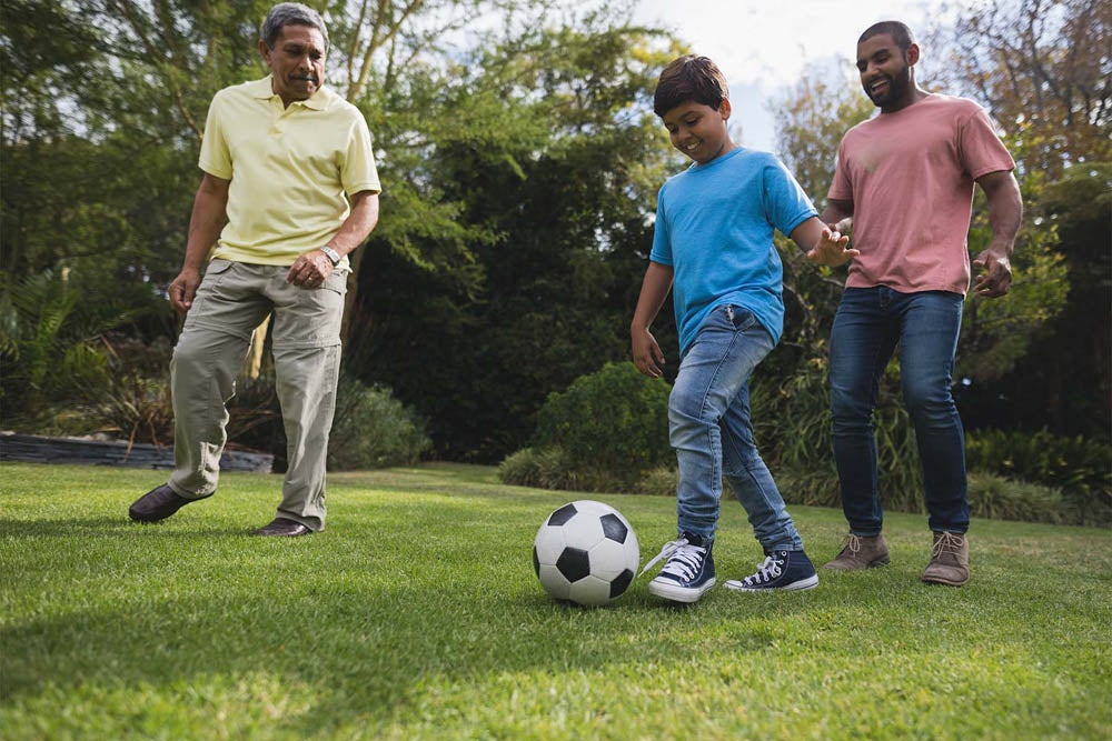 A boy in a blue shirt is playing soccer on a grassy lawn, while two adults, one in a yellow shirt and the other in a pink shirt, watch and interact with him.