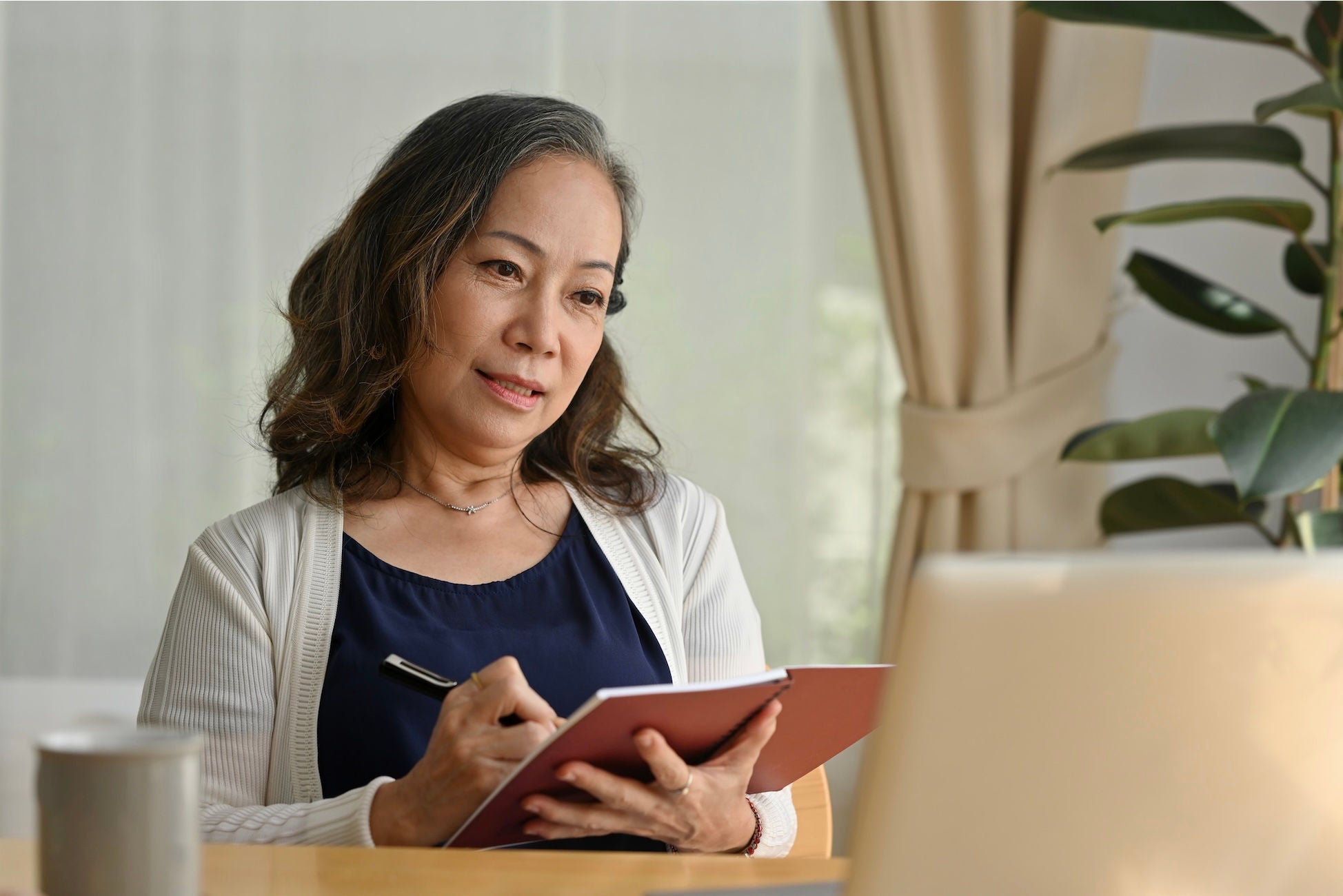 Woman smiling and looking at a laptop screen while taking notes in a notebook.